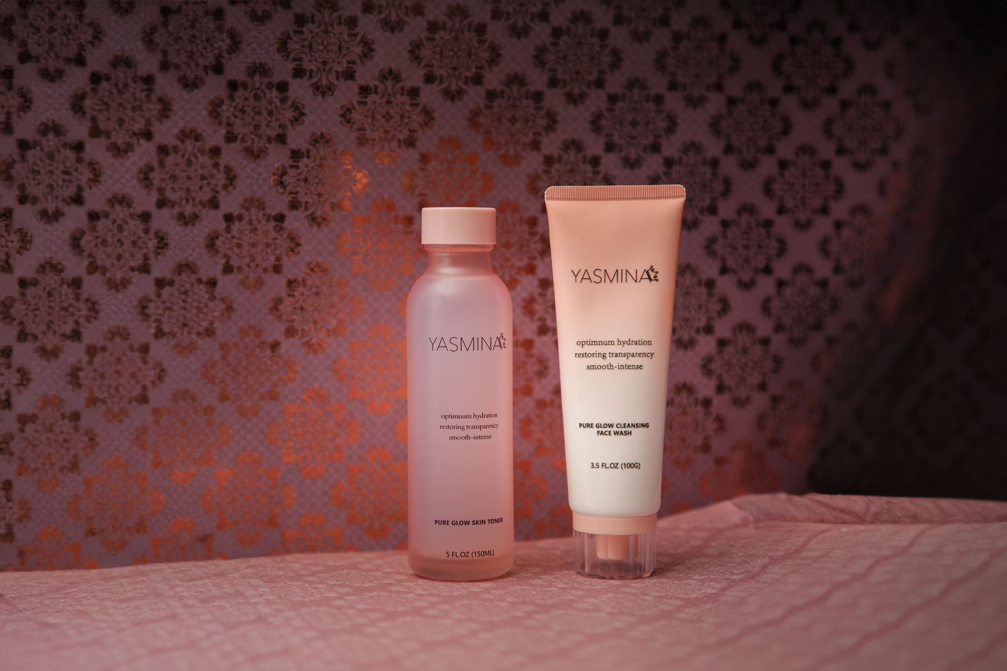 Zwei Yasmina-Hautpflegeprodukte vor gemustertem Hintergrund, ein Toner und ein Cleanser in Rosa.