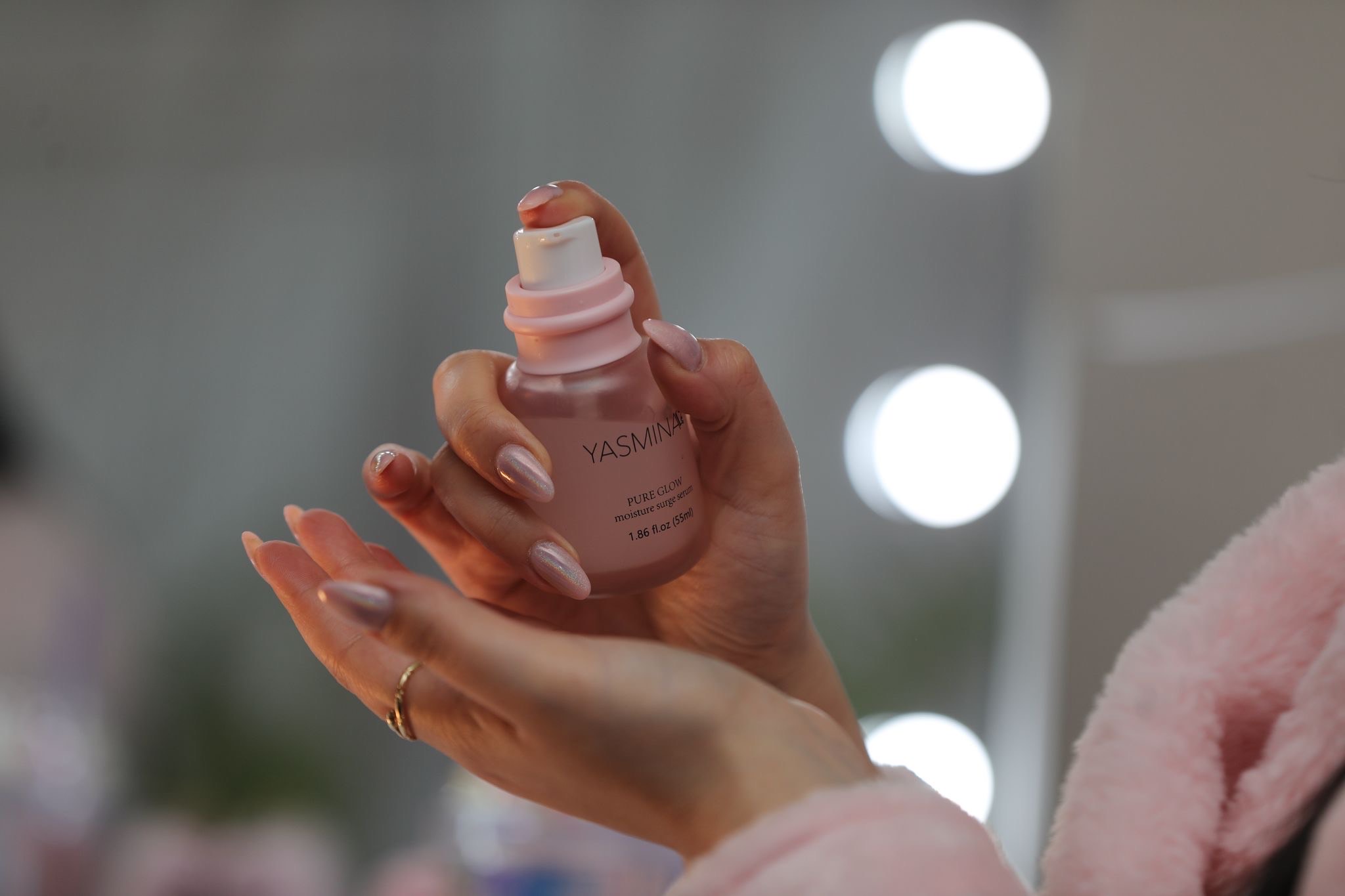 Hand hält rosa Kosmetikflasche vor Spiegel mit Lichtern, pastellrosa Maniküre.